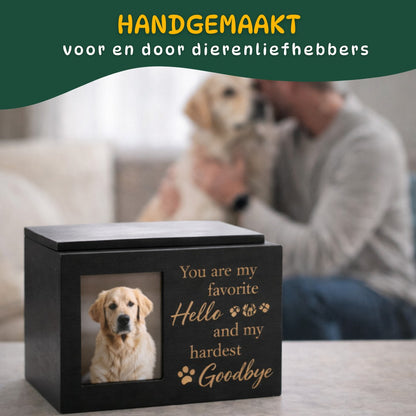 Hondenurn met fotovak en schuifdeksel op de voorgrond, met eigenaar en hond onscherp op de achtergrond als symbool van herinnering en liefde